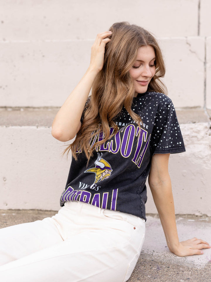 A woman wearing a vintage Minnesota Vikings black graphic t-shirt and white pants, sitting against a light concrete wall.