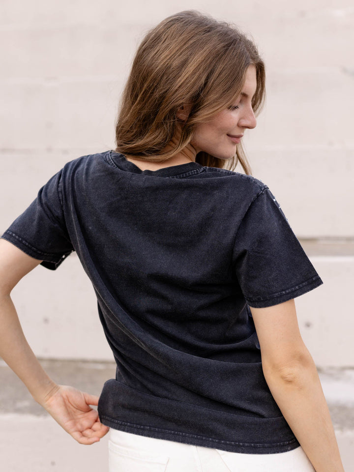 A woman wearing a faded black short-sleeve t-shirt, shown from the back as she looks over her shoulder against a light-colored brick wall.