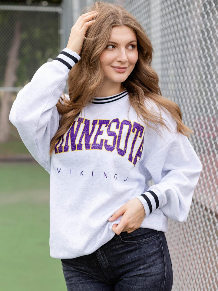 A woman wearing a white Minnesota Vikings sweatshirt with purple and gold lettering, paired with dark jeans, standing in front of a chain-link fence.