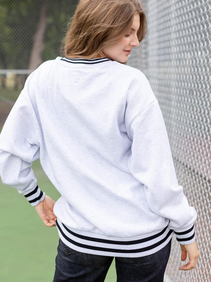 A woman wearing a white crewneck sweatshirt with black striped ribbing on the collar, cuffs, and hem, shown from the back in an outdoor setting next to a chain-link fence.