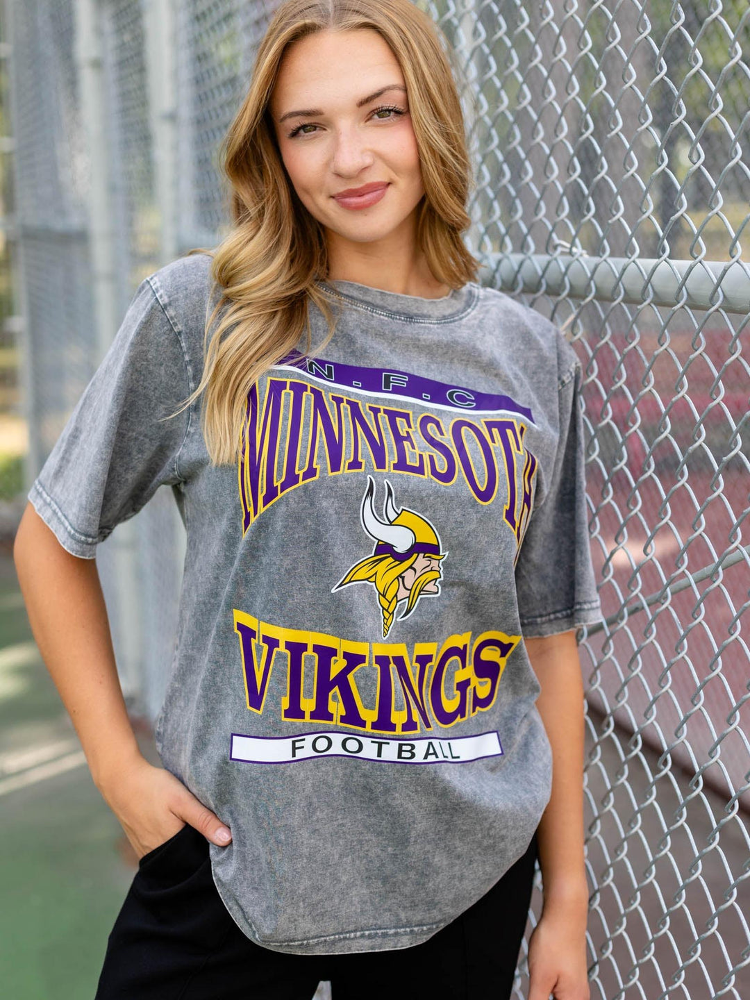 A woman wearing a gray Minnesota Vikings football graphic t-shirt, standing in front of a chain-link fence.