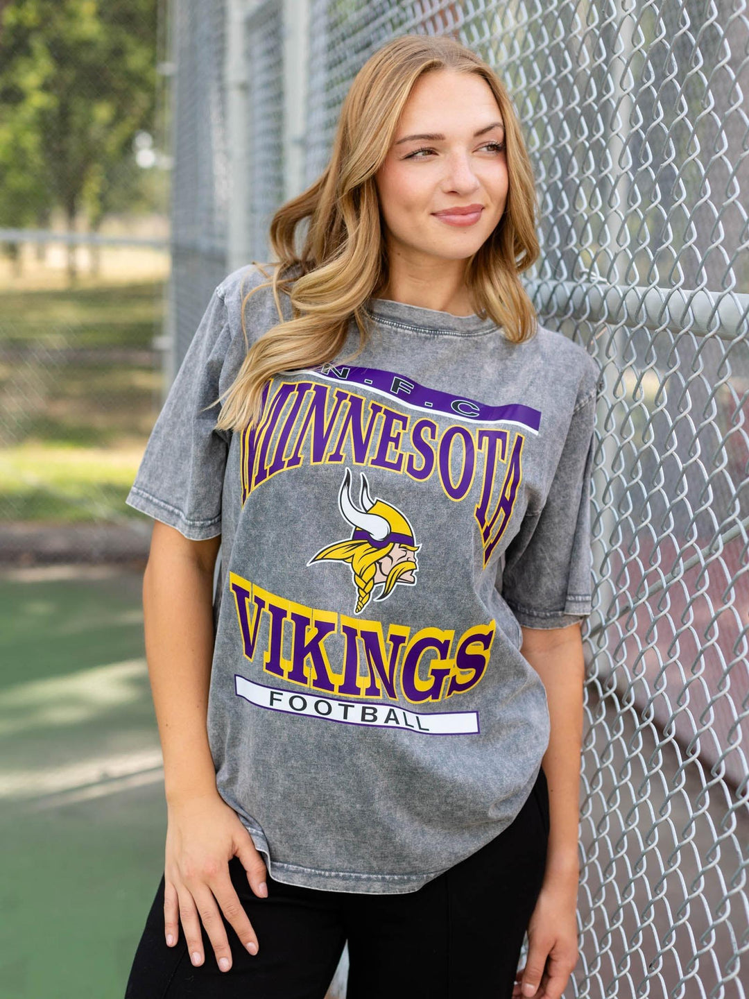 A woman wearing a gray Minnesota Vikings Football graphic t-shirt stands in front of a chain-link fence.