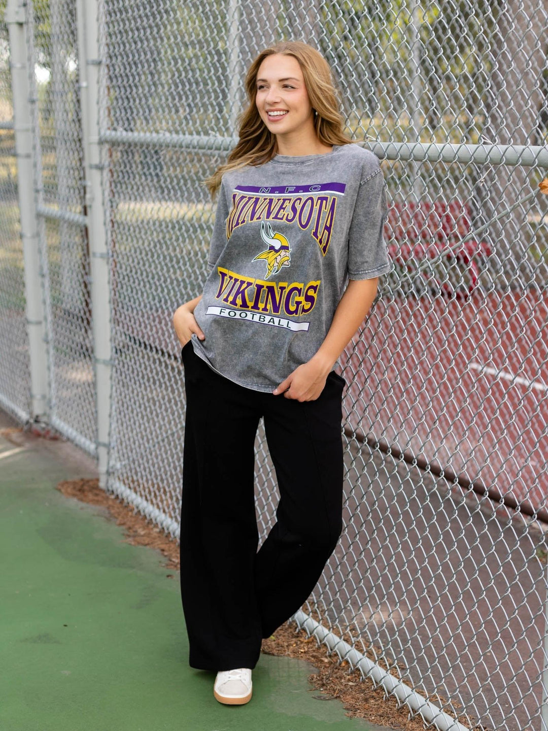 A woman stands outdoors in front of a chain-link fence wearing a gray Minnesota Vikings graphic t-shirt, black wide-leg pants, and white sneakers.