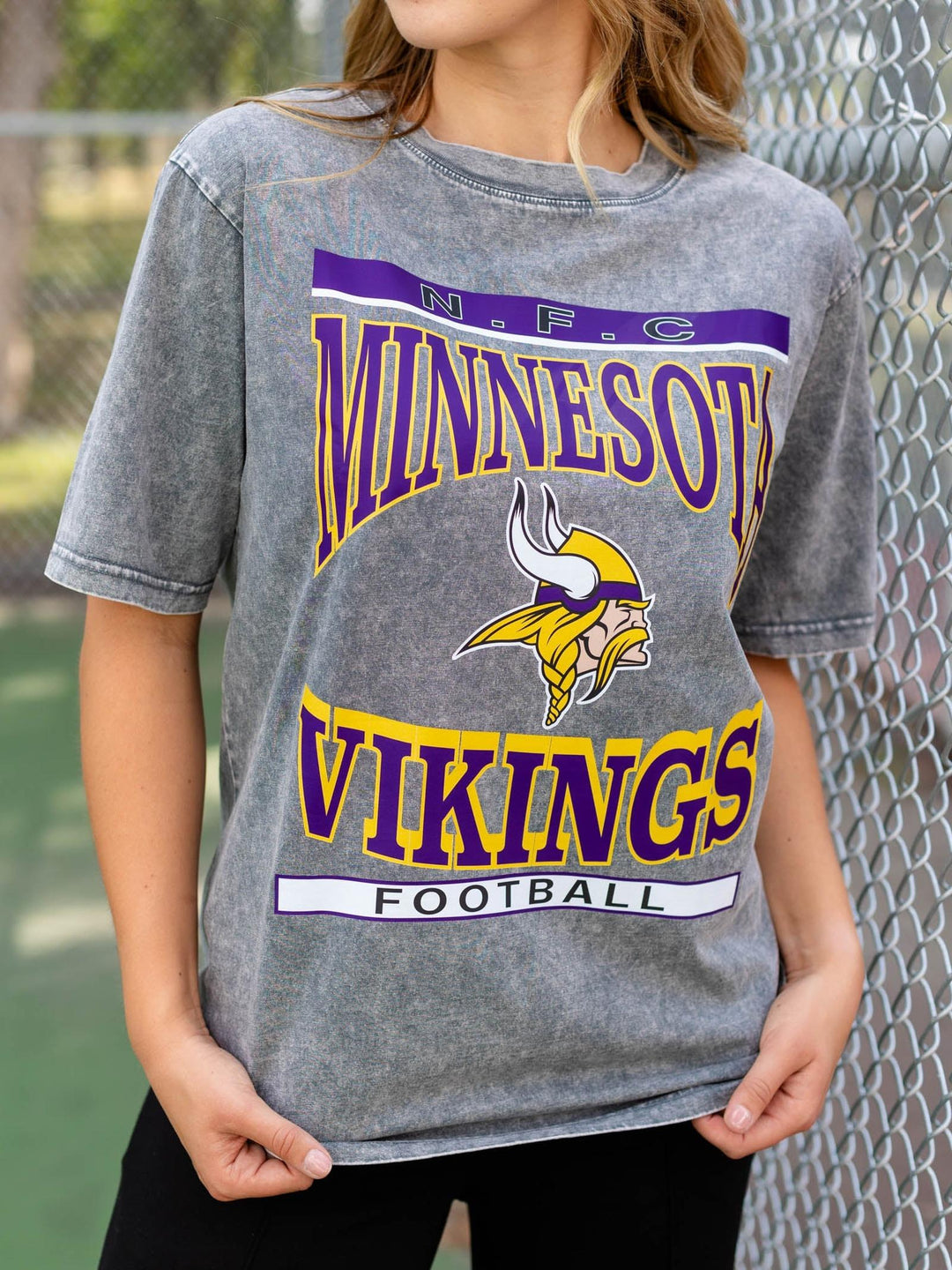 A person wearing a gray mineral-wash graphic t-shirt featuring the Minnesota Vikings logo and text, standing outdoors in front of a chain-link fence.