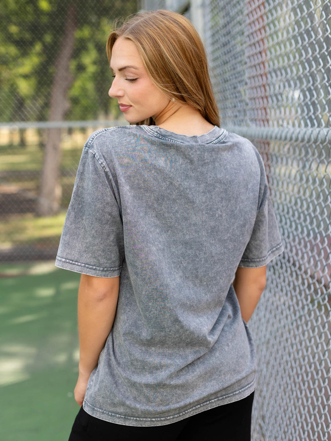 A woman wearing a gray acid-wash t-shirt, shown from the back in an outdoor setting with a chain-link fence and green court visible in the background.
