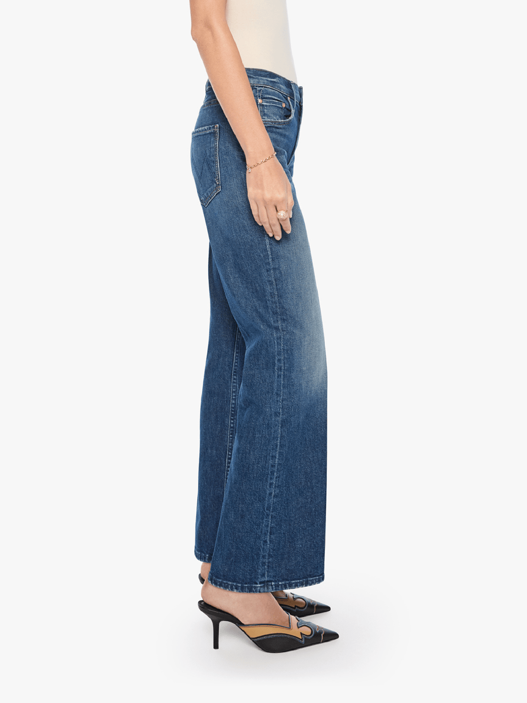 A side profile view of a model wearing blue wide-leg denim jeans, styled with a cream top and black pointed-toe mule heels with gold detailing, against a plain white background.