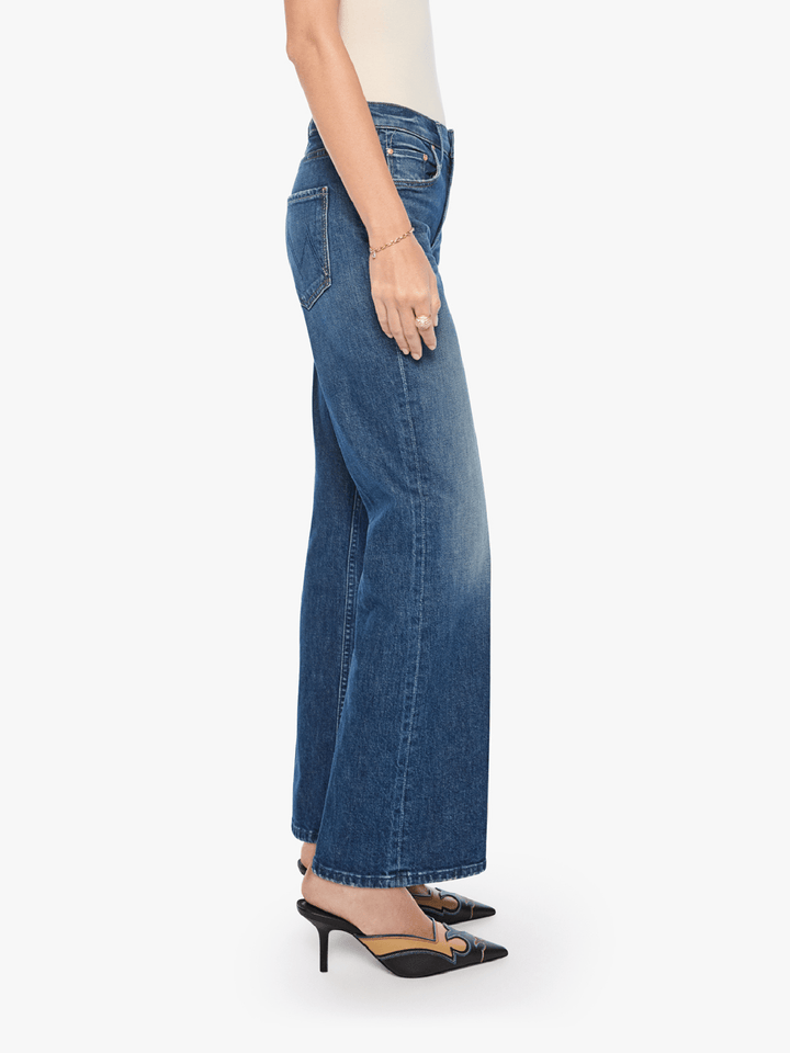 A side profile view of a model wearing blue wide-leg denim jeans, styled with a cream top and black pointed-toe mule heels with gold detailing, against a plain white background.