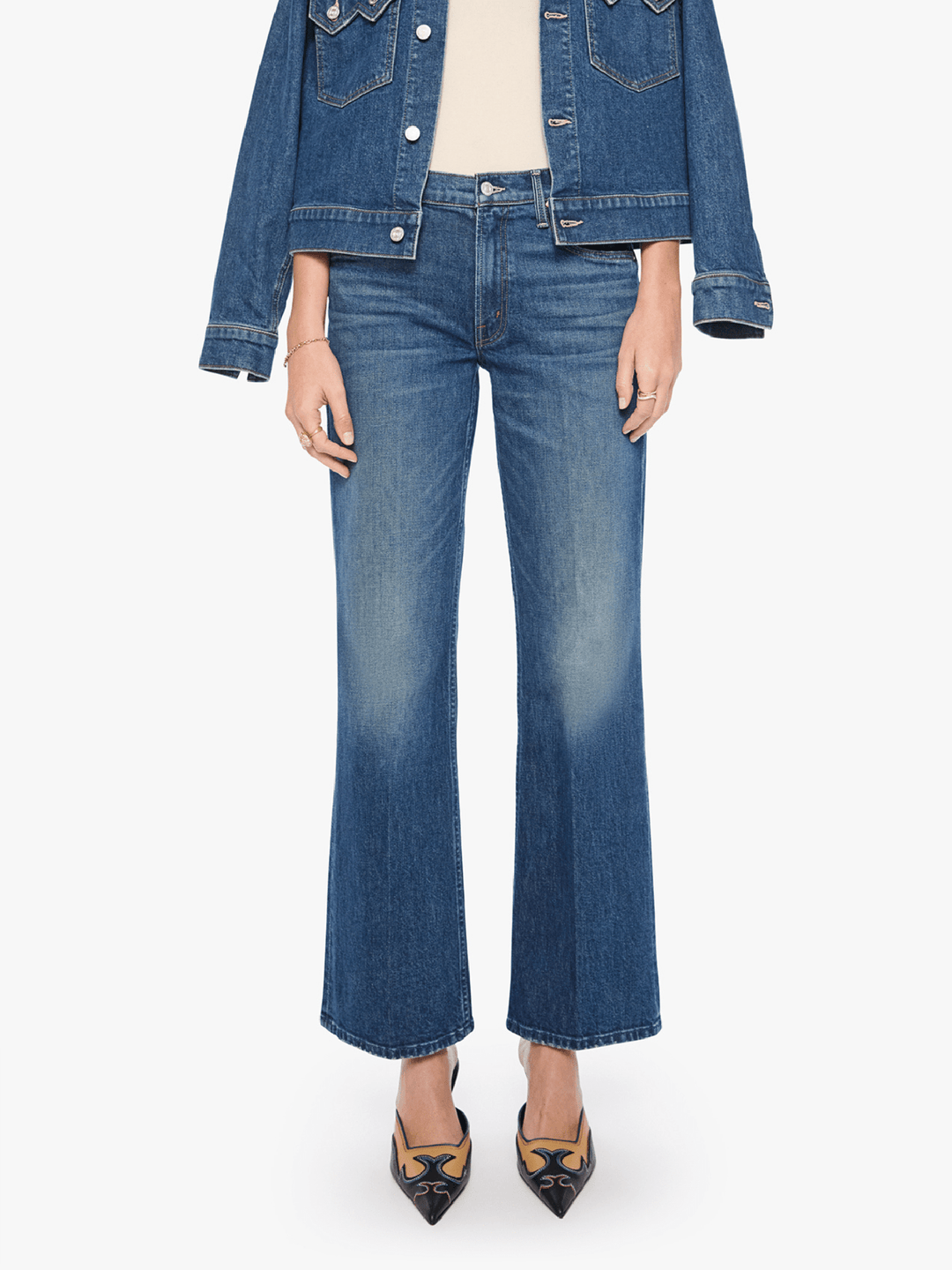 A model wearing a pair of medium-wash blue denim flare jeans, styled with a cropped denim jacket, cream top, and black-and-gold pointed-toe heels against a plain white background.