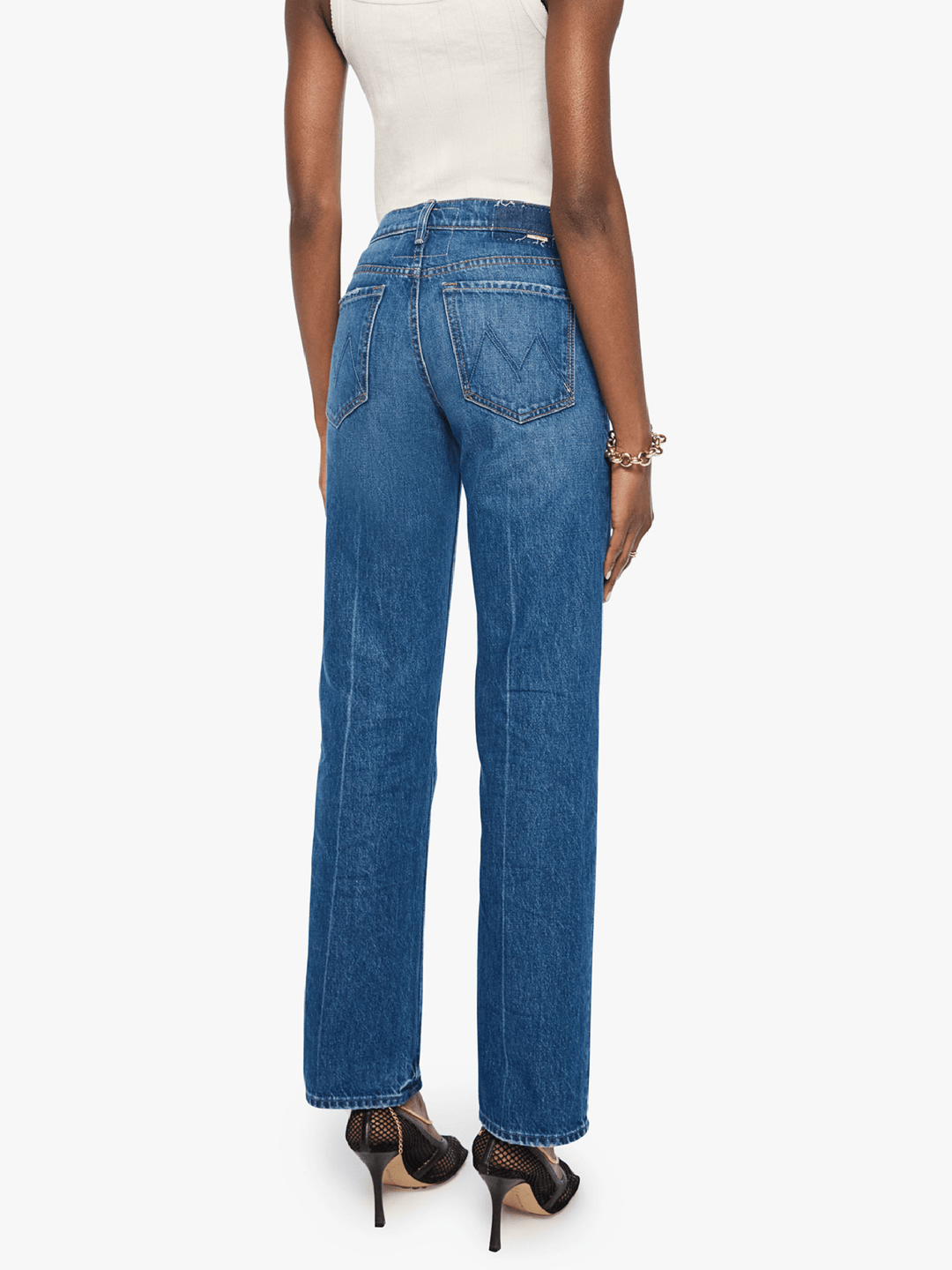 A model shown from the back wearing high-waisted, wide-leg blue denim jeans, a white ribbed tank top, and black high-heeled sandals against a plain white background.