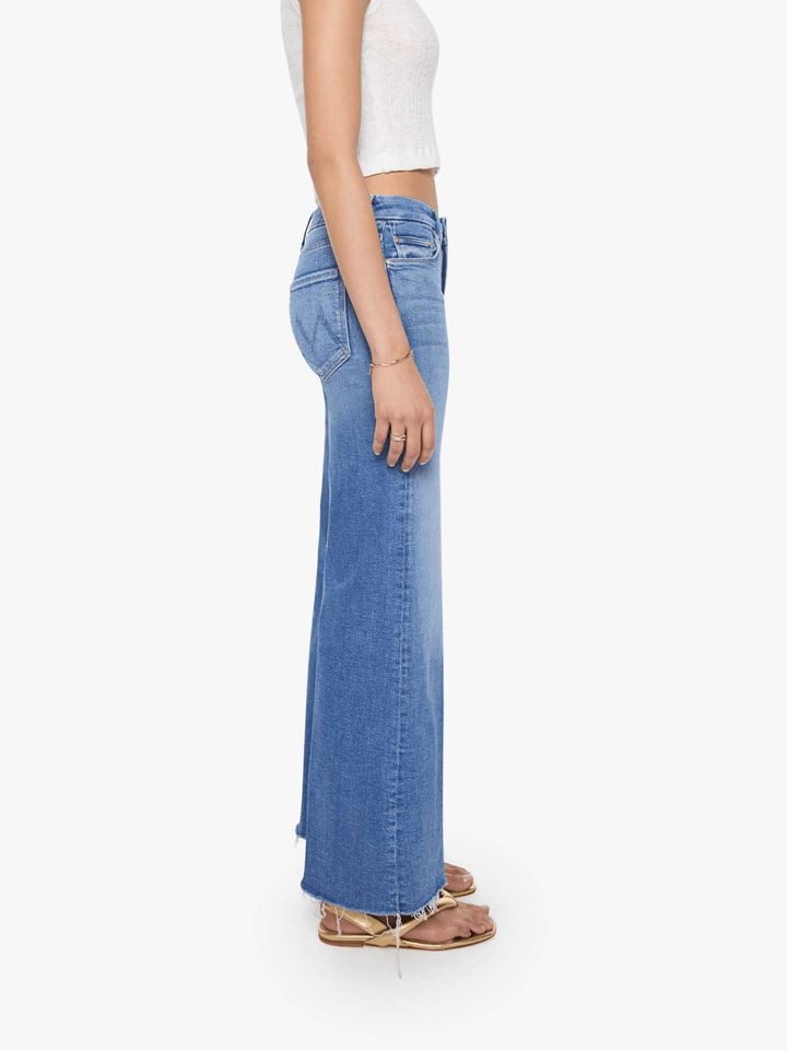 A side profile view of a model wearing high-waisted, wide-leg blue denim jeans, a white cropped tank top, and flat sandals against a plain white background.