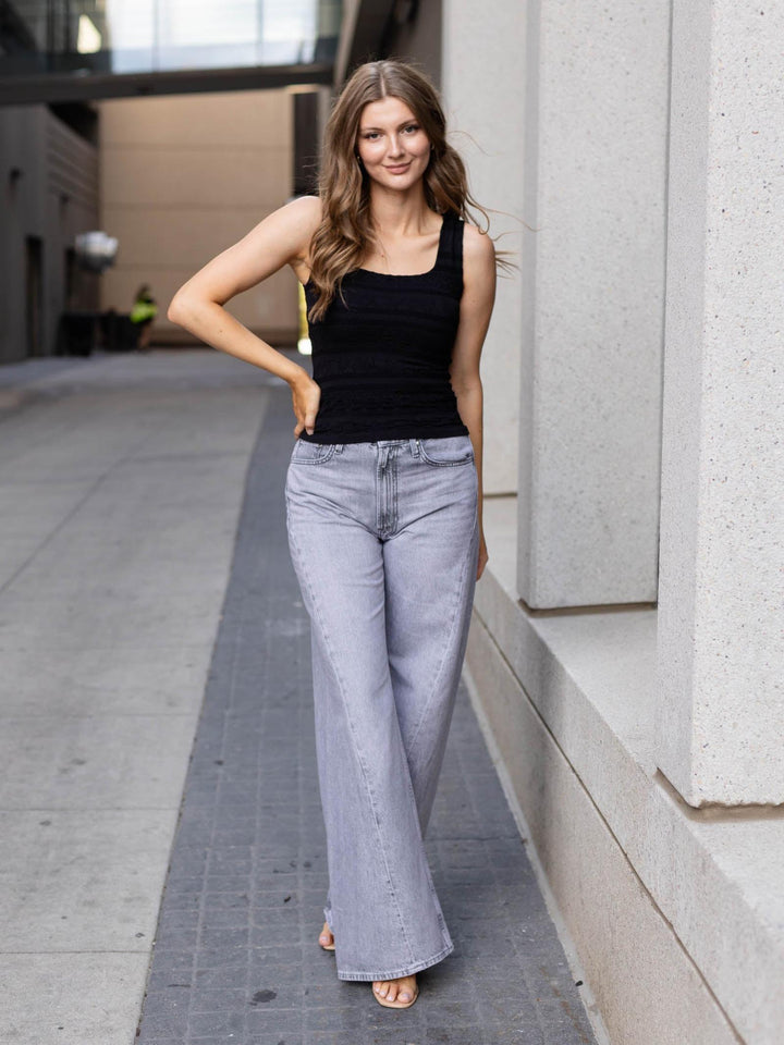 A woman posing outdoors on a city sidewalk, wearing a black ribbed tank top and light-wash wide-leg jeans.