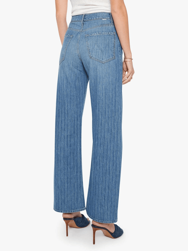 Rear view of a model wearing high-waisted, wide-leg blue denim jeans with a subtle vertical stripe texture, paired with dark blue mule heels against a light gray studio background.