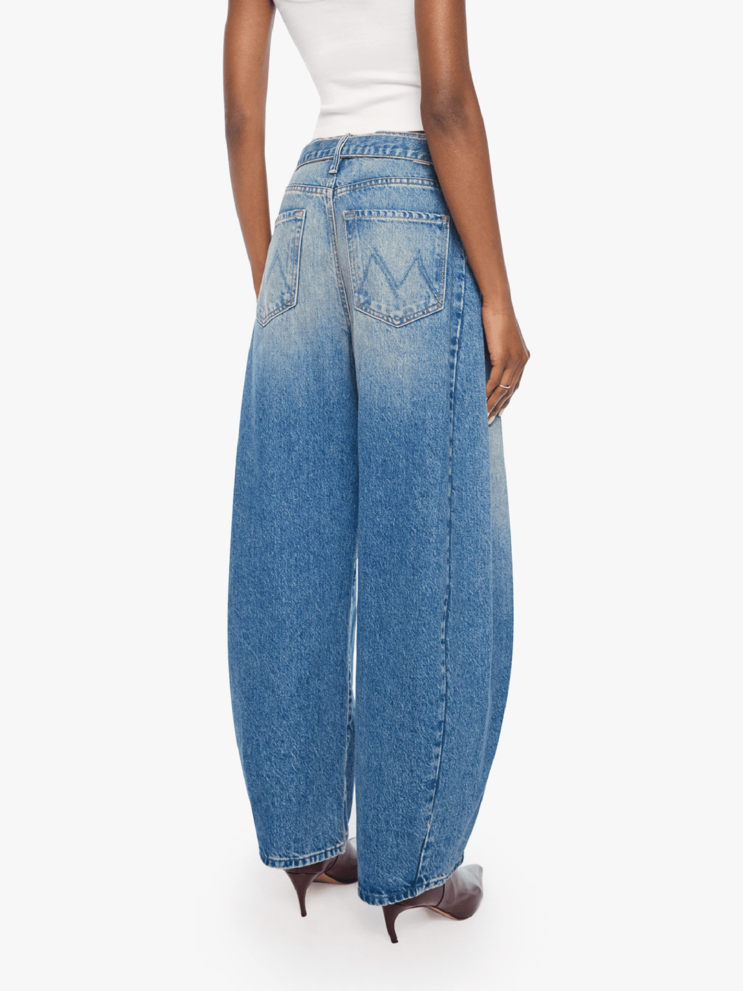 A model shown from the back wearing high-waisted, wide-leg blue denim jeans with a faded wash and decorative stitching on the back pockets, paired with a white top and brown heels.