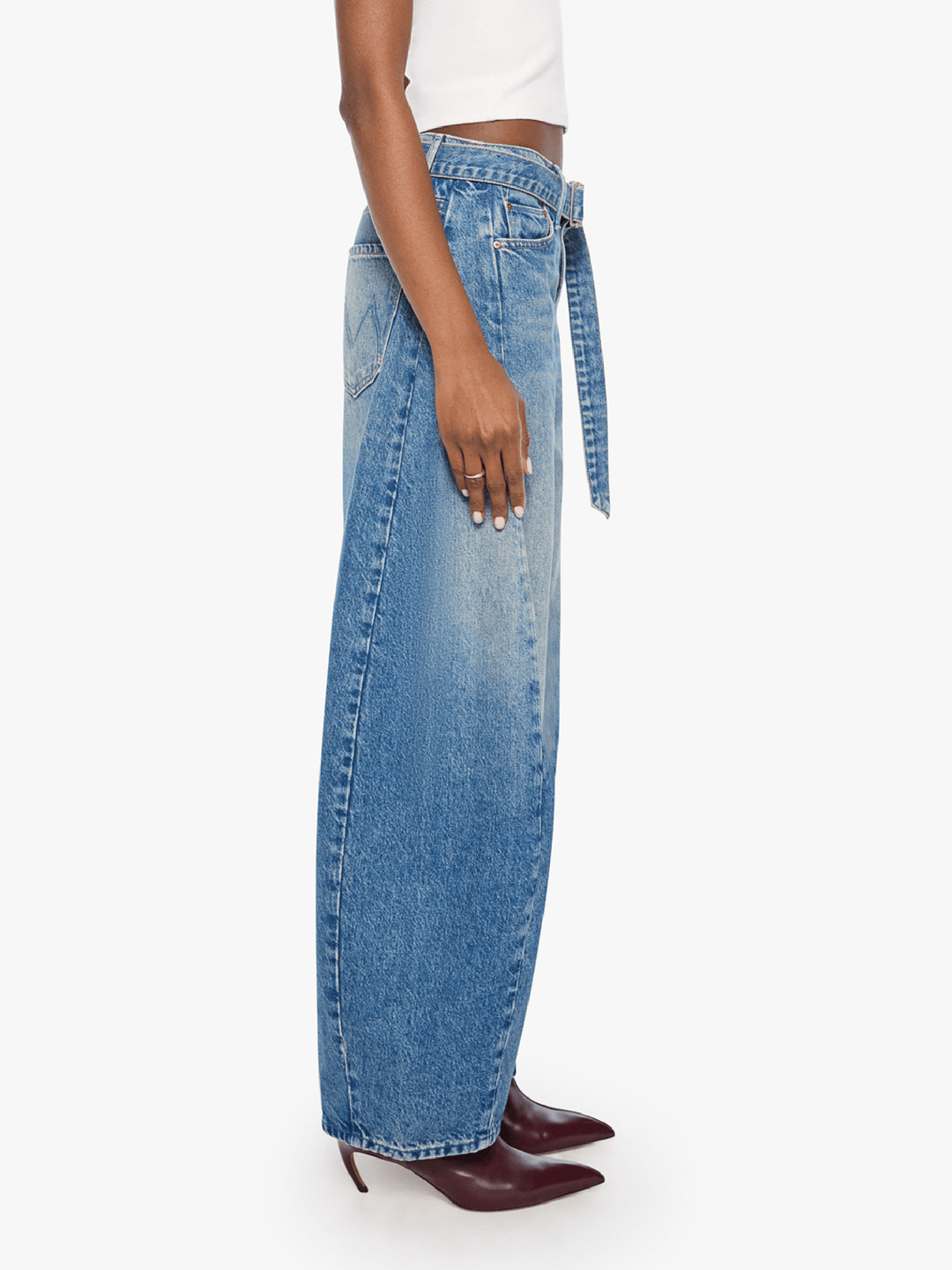 A side-profile view of a model wearing high-waisted, wide-leg blue denim jeans with a matching tie belt, paired with a white crop top and dark burgundy pointed-toe heels, against a plain white background.