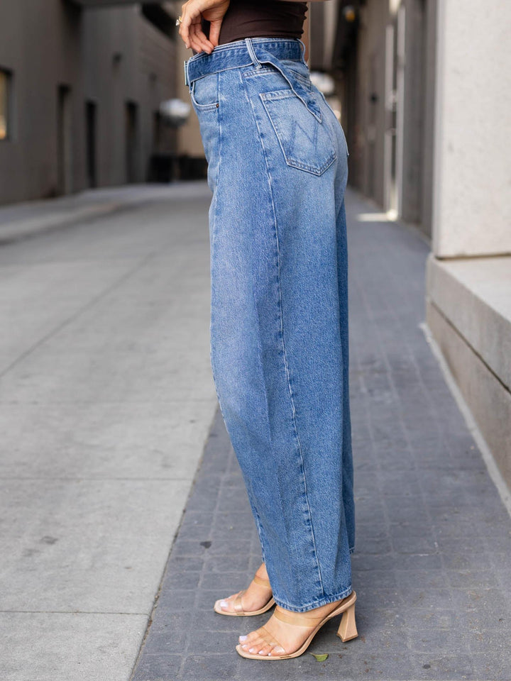 A person wearing wide-leg light blue denim jeans and clear heeled sandals, standing on a city sidewalk between buildings.