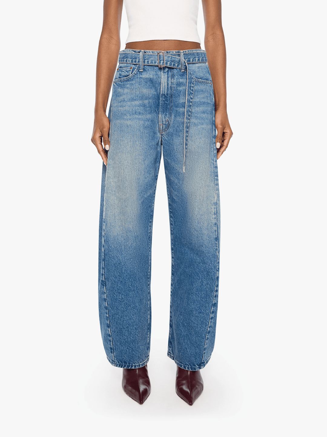 A model wearing a pair of wide-leg, high-waisted blue denim jeans with a faded wash, paired with a white crop top and dark brown pointed-toe boots, shot against a plain white studio background.