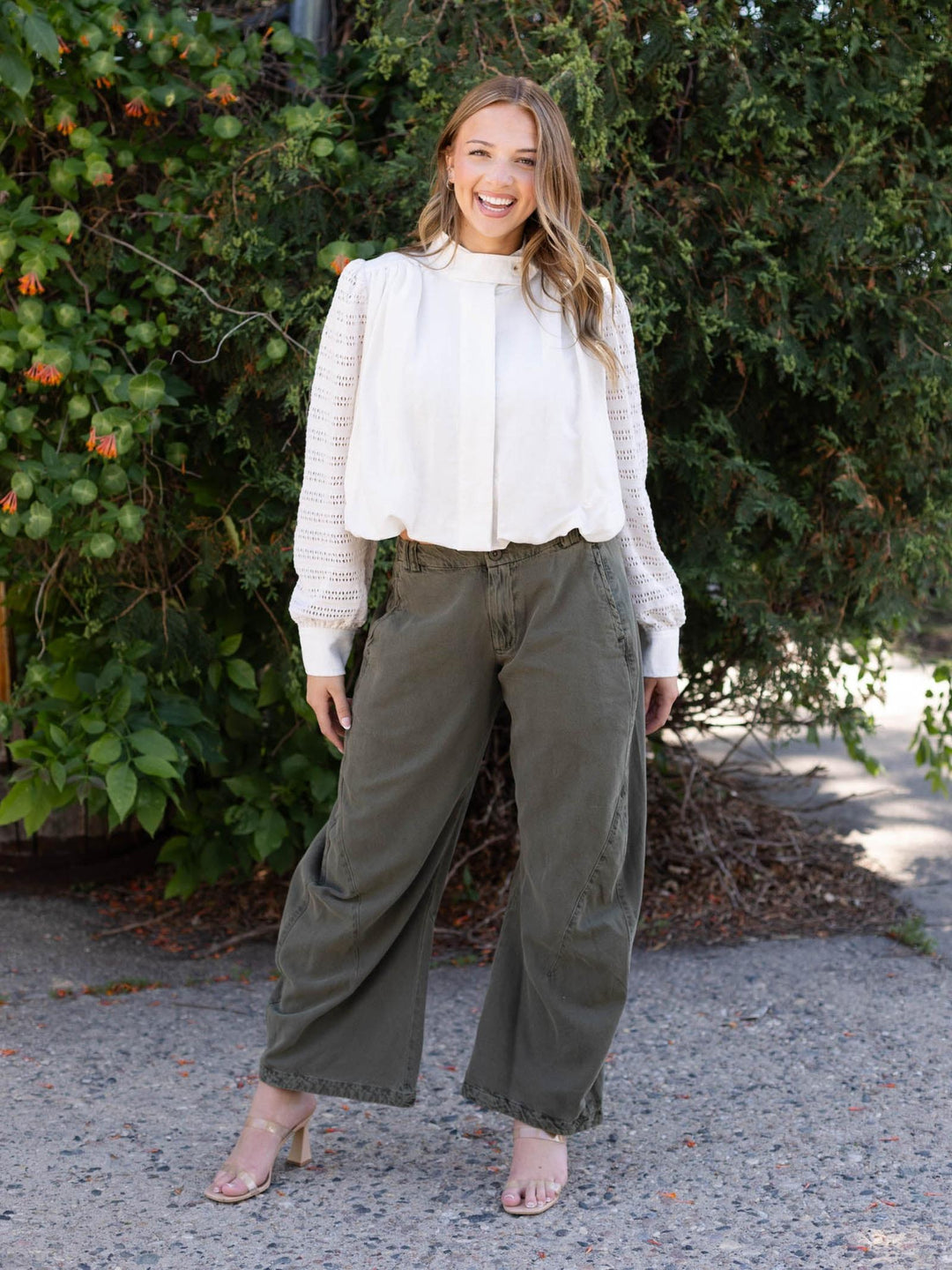 Woman smiling in an Off White Mystree Bubble Hem Jacket with crochet knit sleeves and olive wide-leg pants.