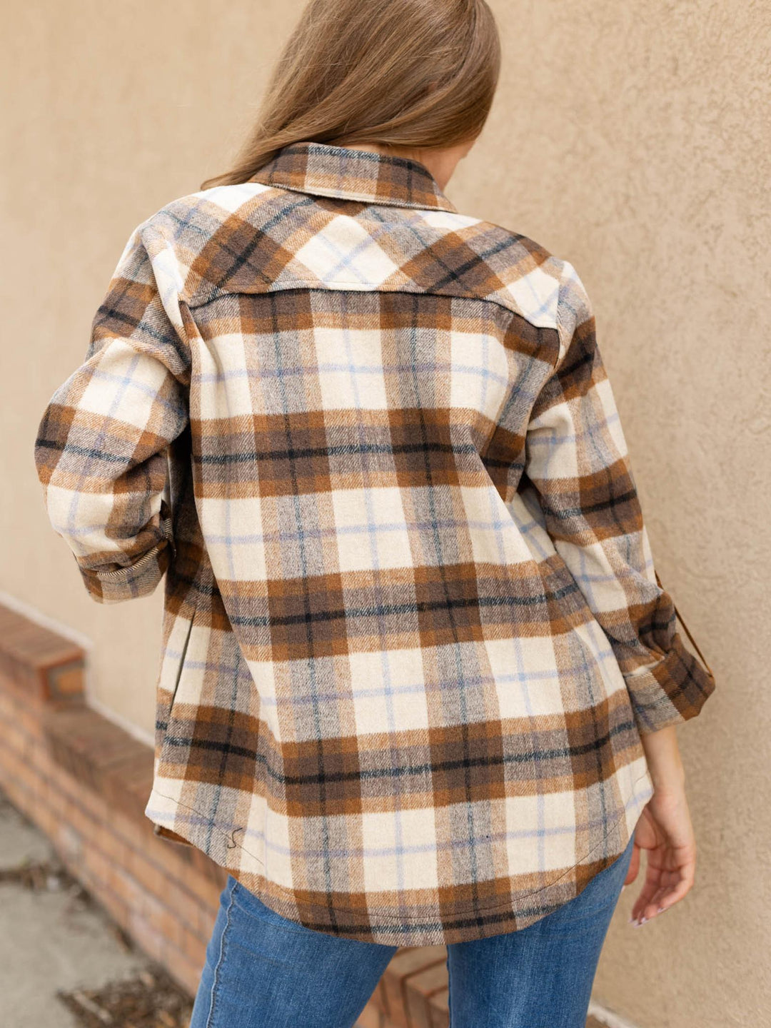 A woman viewed from behind wearing a brown, beige, and blue plaid flannel shirt with rolled-up sleeves, paired with blue jeans, standing against a beige stucco wall.
