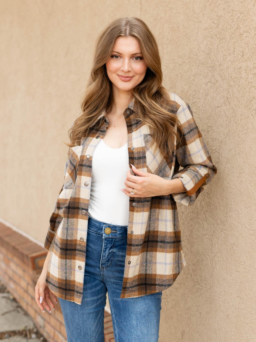 A woman wearing a brown, beige, and white plaid button-up shirt over a white tank top and blue jeans, standing against a textured beige stucco wall.
