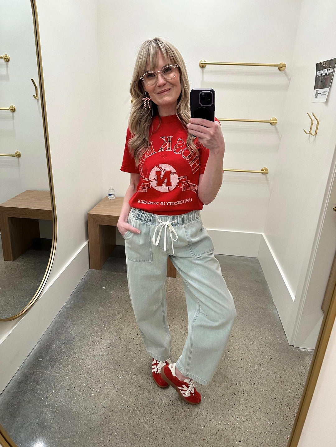 Person taking a mirror selfie wearing a red t-shirt and light gray pants in a fitting room.