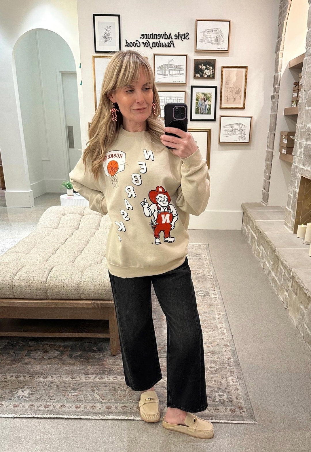 Woman taking a mirror selfie wearing a beige sweatshirt with graphics and text, standing in a room with a couch and framed pictures on the wall.