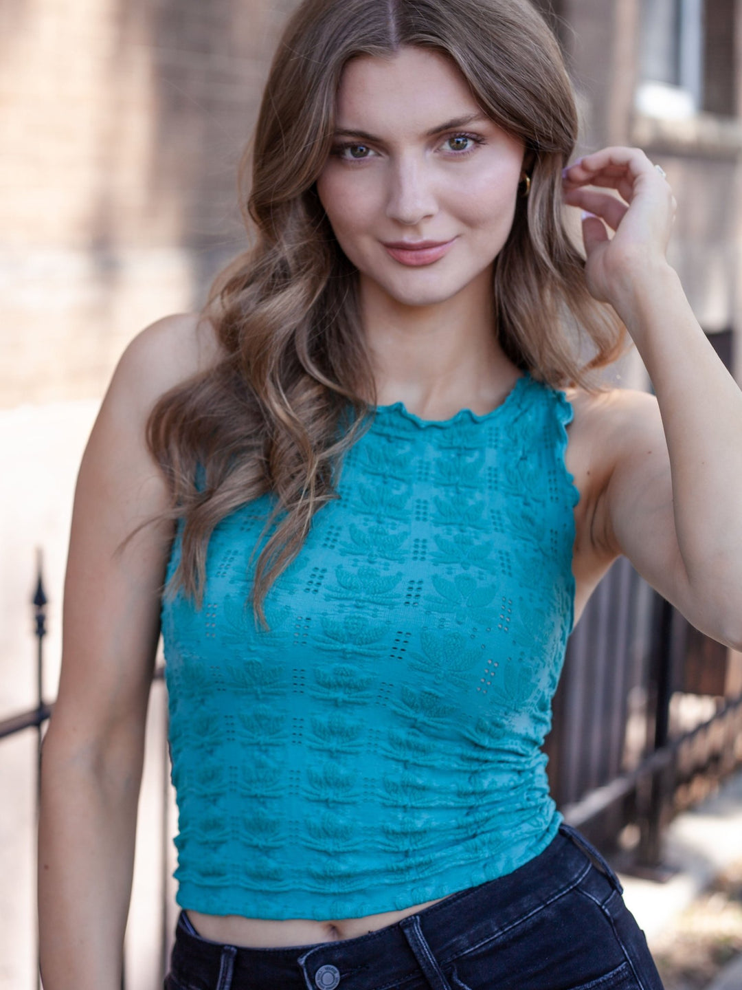 A woman wearing a sleeveless, textured turquoise blue crop top and dark denim jeans, standing outdoors against a blurred brick wall background.