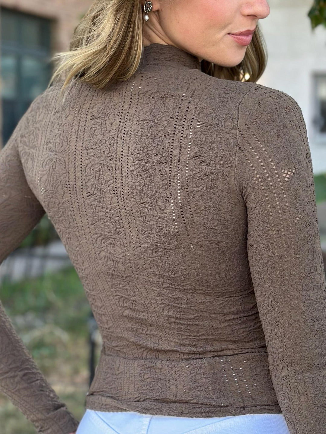 A woman wearing a brown long-sleeve mock neck top with intricate textured knit patterns, shown from a rear three-quarter view.