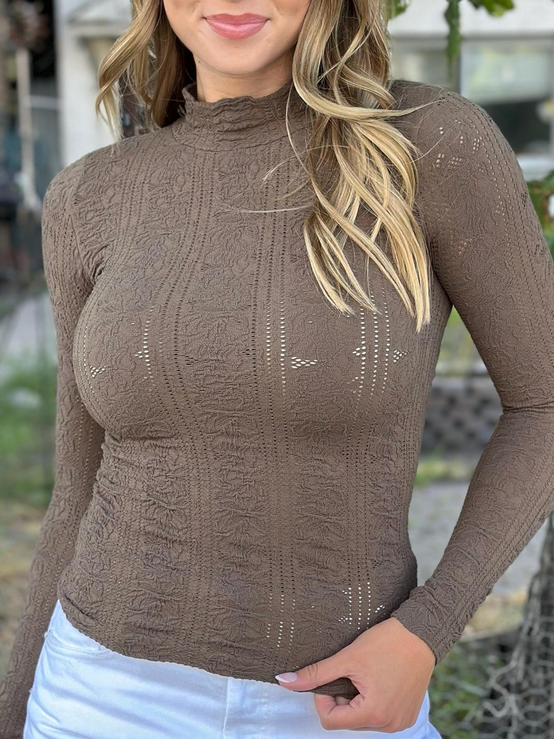 A woman wearing a brown long-sleeve mock neck knit top with textured knit patterns and small cutout details, paired with white pants.