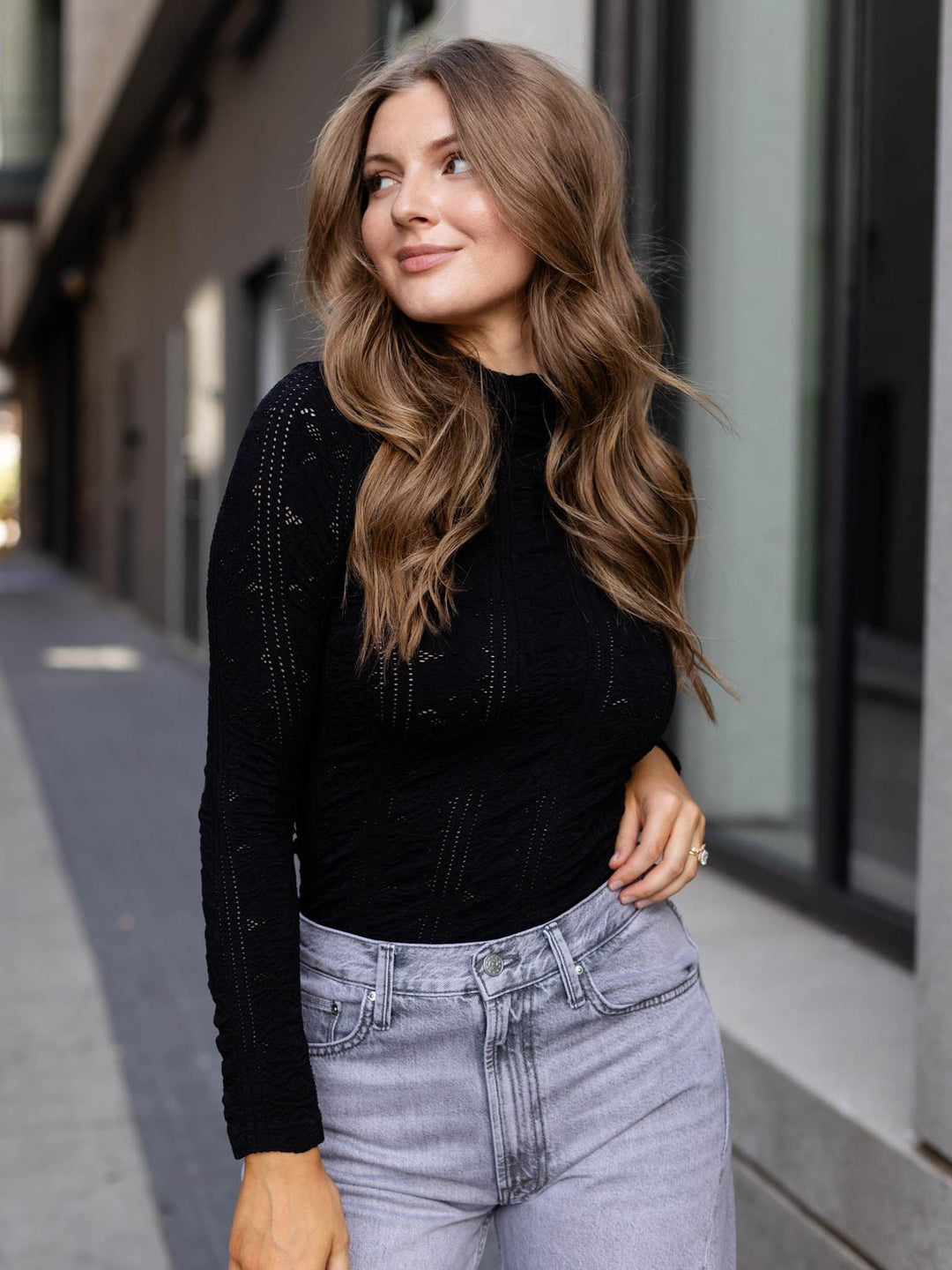 A woman with long wavy light brown hair wearing a black long-sleeve knit top and light-wash gray jeans, standing outdoors on a city sidewalk.