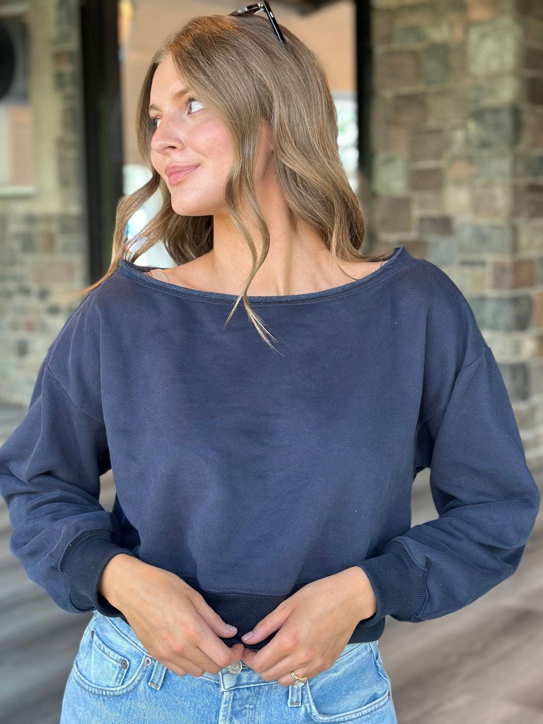 A woman wearing a navy blue wide-neck long-sleeve sweatshirt and light blue jeans, standing in front of a brick building.