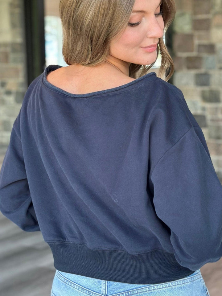 A woman wearing a navy blue off-the-shoulder sweatshirt, shown from the back. She is wearing light wash blue jeans, and the background features a blurred brick wall.
