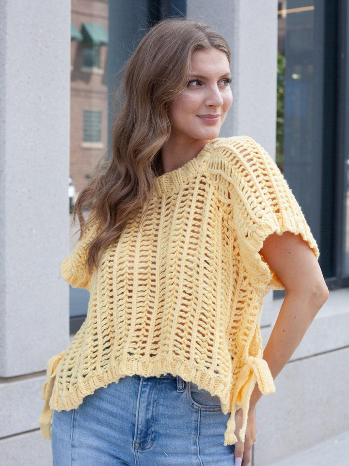 A woman wearing a pale yellow open-knit short-sleeve top with side tie details and blue jeans, standing outdoors against a light gray stone wall.