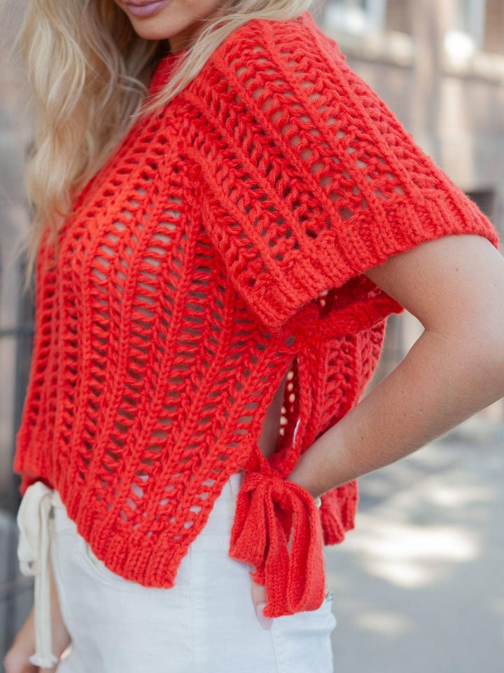 A woman wearing a bright coral-red open-knit short-sleeve shrug or poncho with tie details at the back, paired with white pants, photographed outdoors.