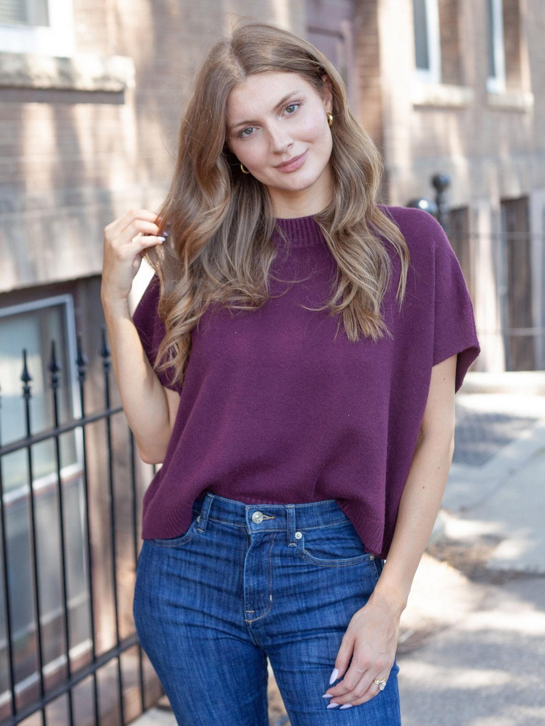 A woman wearing a short-sleeve, crewneck knit sweater in a deep plum purple, paired with blue jeans, standing outdoors on a city sidewalk.