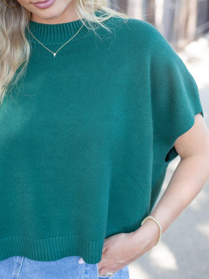 A close-up of a woman wearing a dark green short-sleeve knit sweater with a ribbed crew neck and hem, paired with light blue jeans.