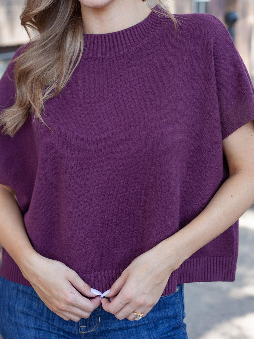 A woman wearing a short-sleeve crewneck knit sweater in a deep plum purple, paired with blue jeans.