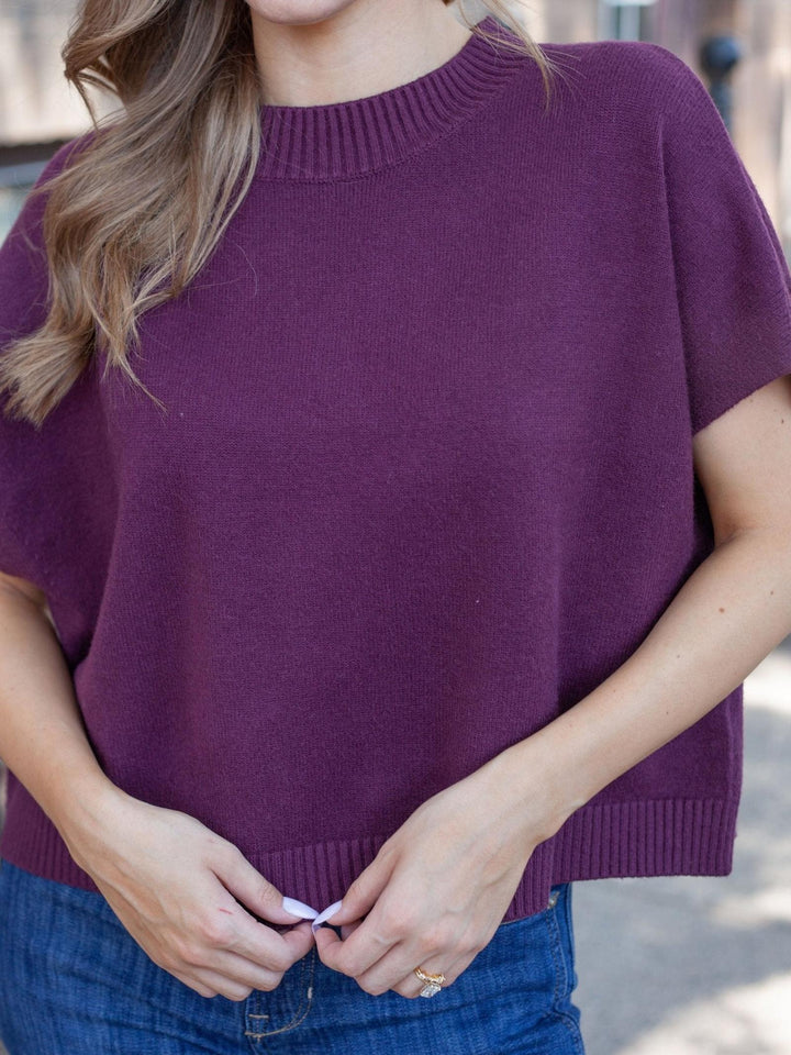 A woman wearing a short-sleeve crewneck knit sweater in a deep plum purple, paired with blue jeans.