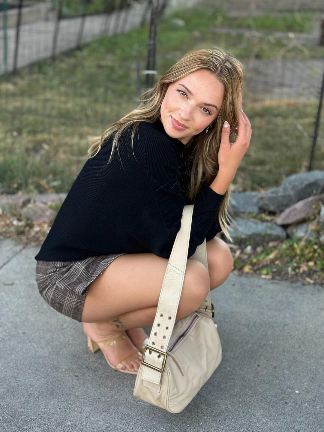 A woman crouching outdoors on a paved path wearing a black long-sleeve top, a brown plaid mini skirt, and beige heeled sandals, while carrying a beige shoulder bag.