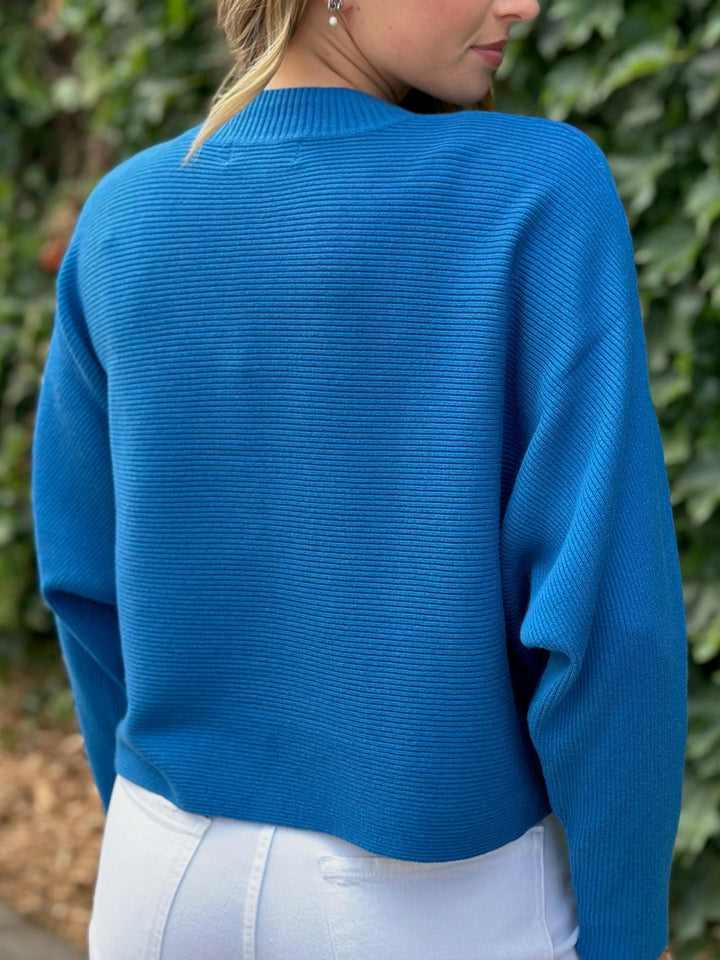 A woman wearing a bright blue ribbed knit sweater, shown from the back. The sweater has a crew neck and long sleeves, paired with white pants, set against a background of green foliage.