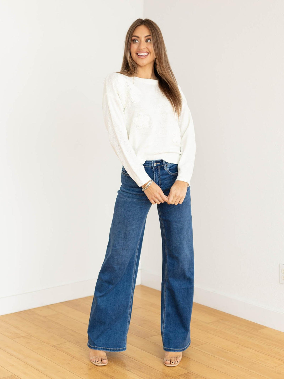 Woman wearing a white sweater and blue jeans standing on a wooden floor with a white wall background