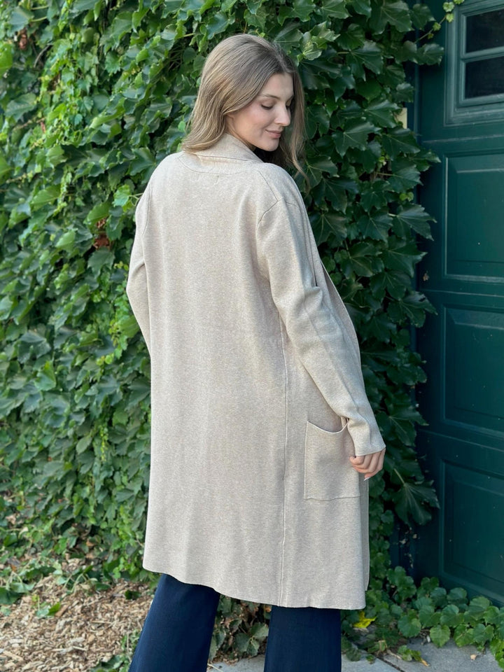 A woman wearing a long, light beige linen-blend cardigan with a mock neck and side pockets, standing outdoors in front of a green ivy-covered wall and dark green door.