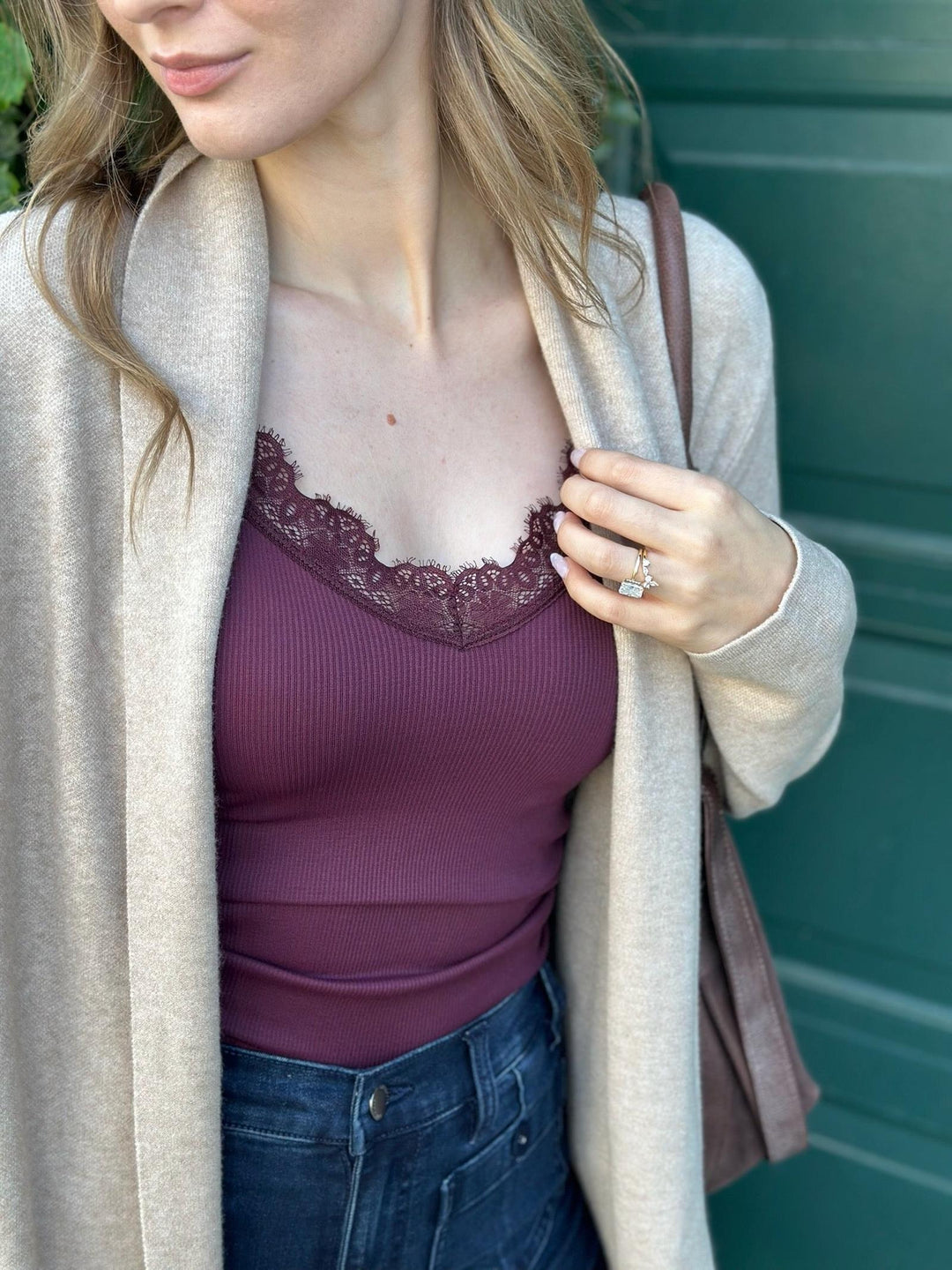 A woman wearing a ribbed plum purple camisole with a lace sweetheart neckline, layered under a light beige cardigan and blue jeans, with a brown shoulder bag.