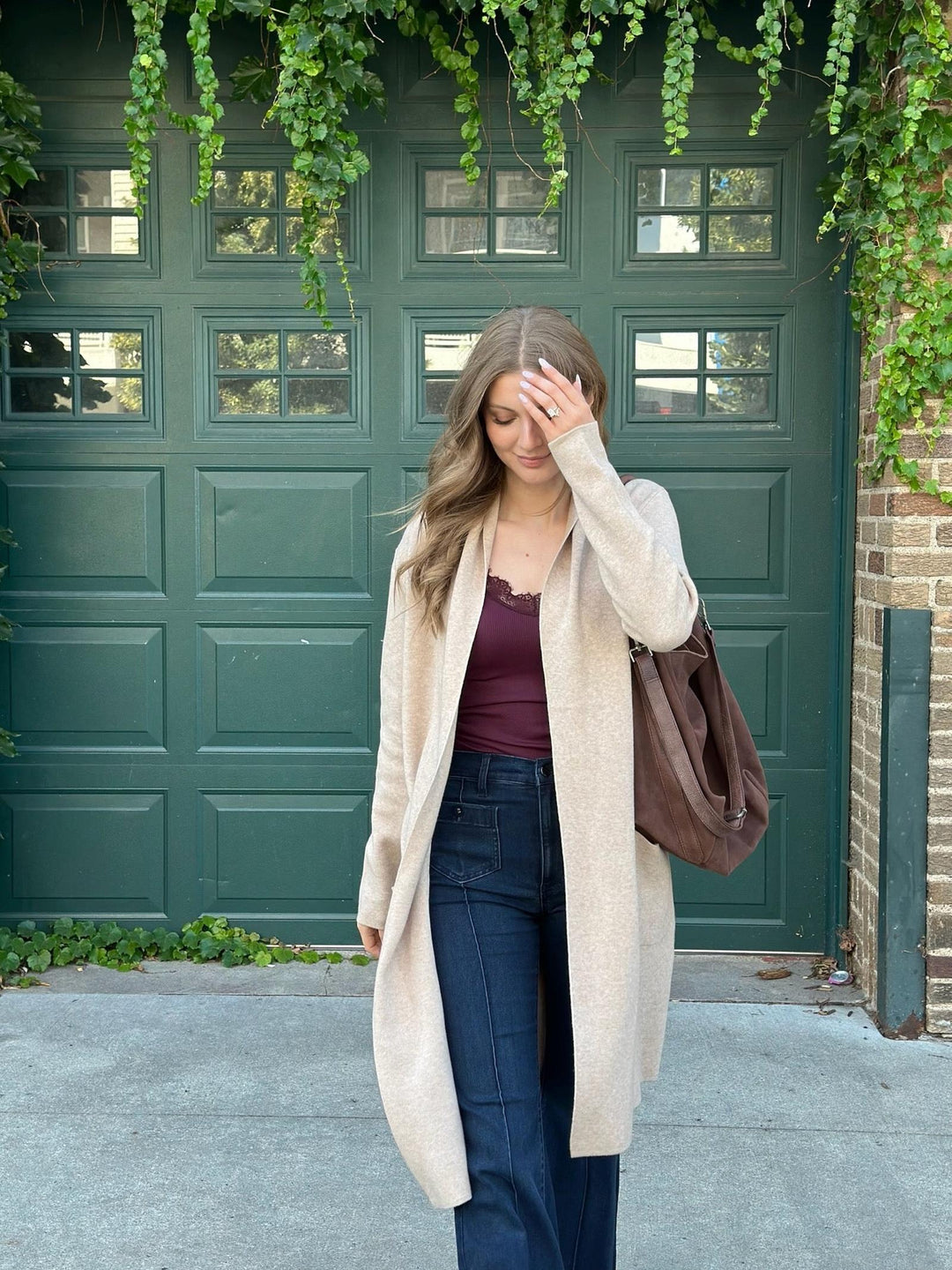 A woman stands in front of a dark green garage door wearing a long cream knit cardigan over a burgundy top and dark blue jeans, carrying a brown shoulder bag.