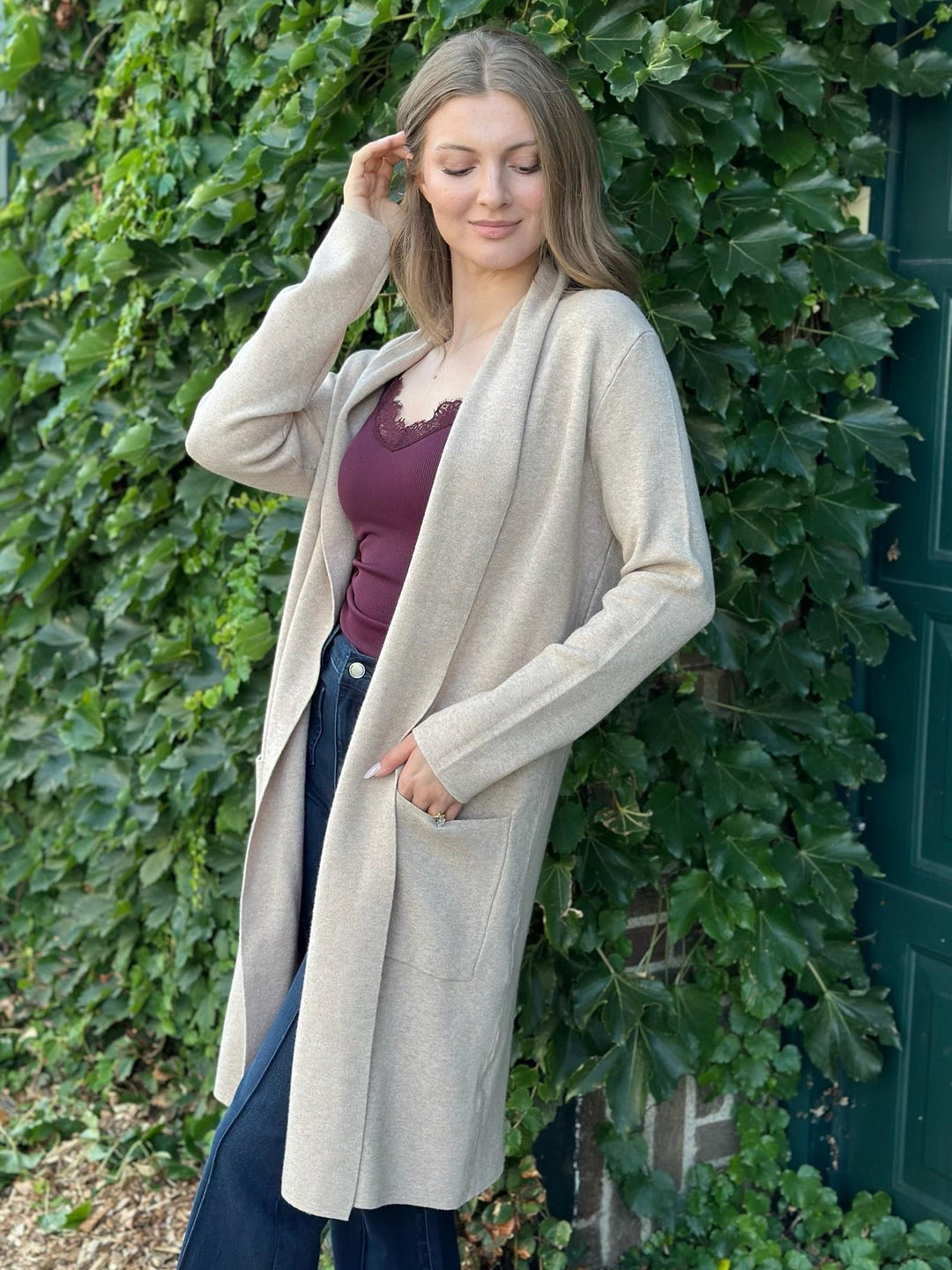 A woman wearing a long, open-front beige cardigan over a burgundy top and dark blue jeans, standing outdoors against a wall covered in green ivy next to a dark green door.