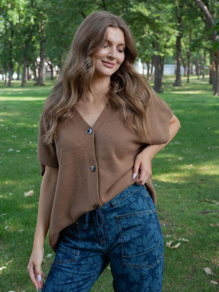 A woman wearing a brown short-sleeve button-front top and blue patterned jeans, standing outdoors in a park with trees and grass in the background.