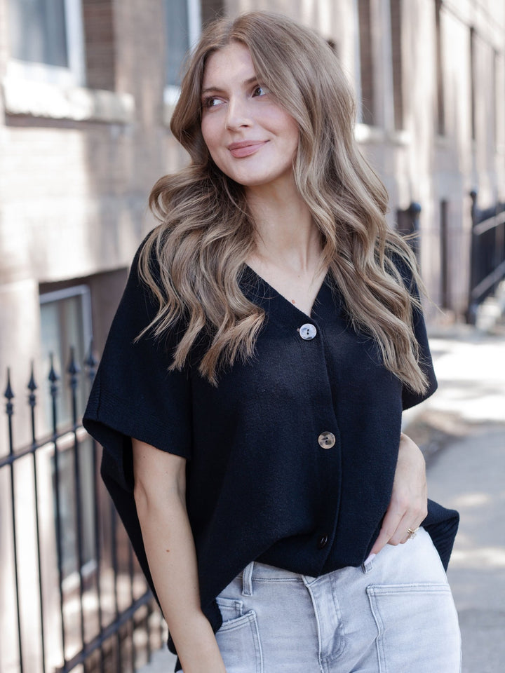 A woman with long wavy light brown hair wearing a black short-sleeve button-up top and light-wash jeans, standing outdoors on a city sidewalk.