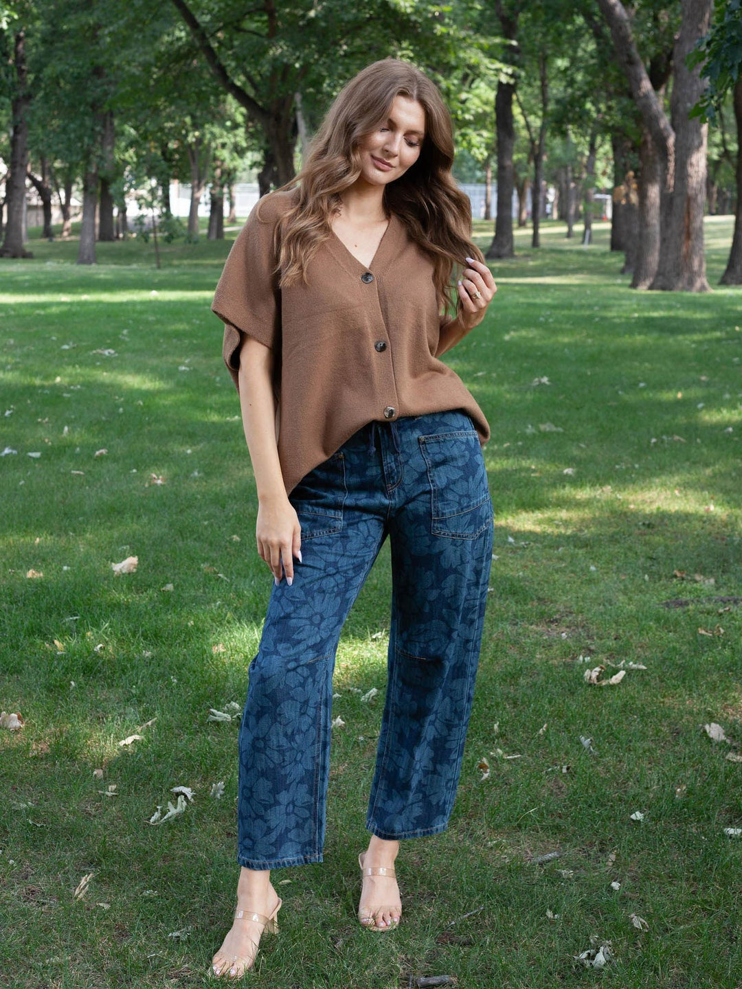 A woman standing barefoot on a grassy park lawn wearing a brown short-sleeve button-front top and blue patterned wide-leg jeans.