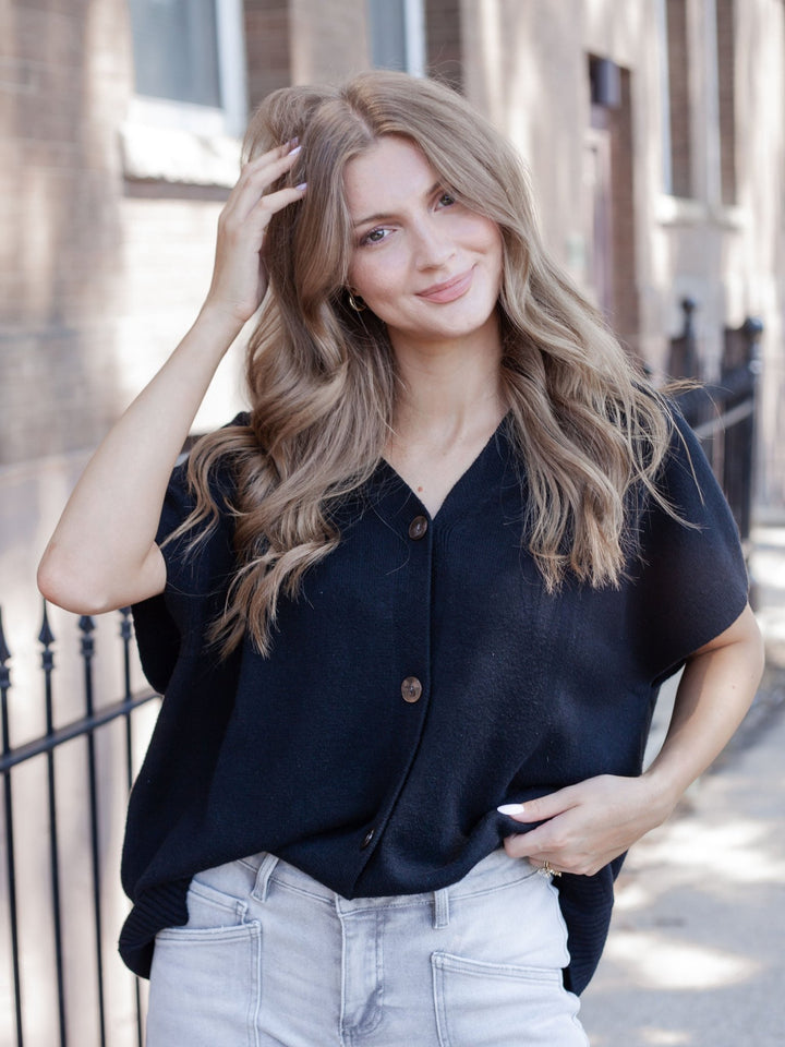 A woman with long wavy blonde hair wearing a black short-sleeve button-up top and light-wash jeans, standing outdoors on a city sidewalk.