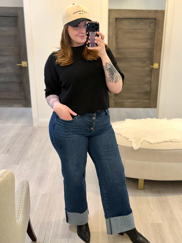 Woman in black PINCH Short Sleeve Sweater Top, dark wash jeans with rolled cuffs, and black boots.