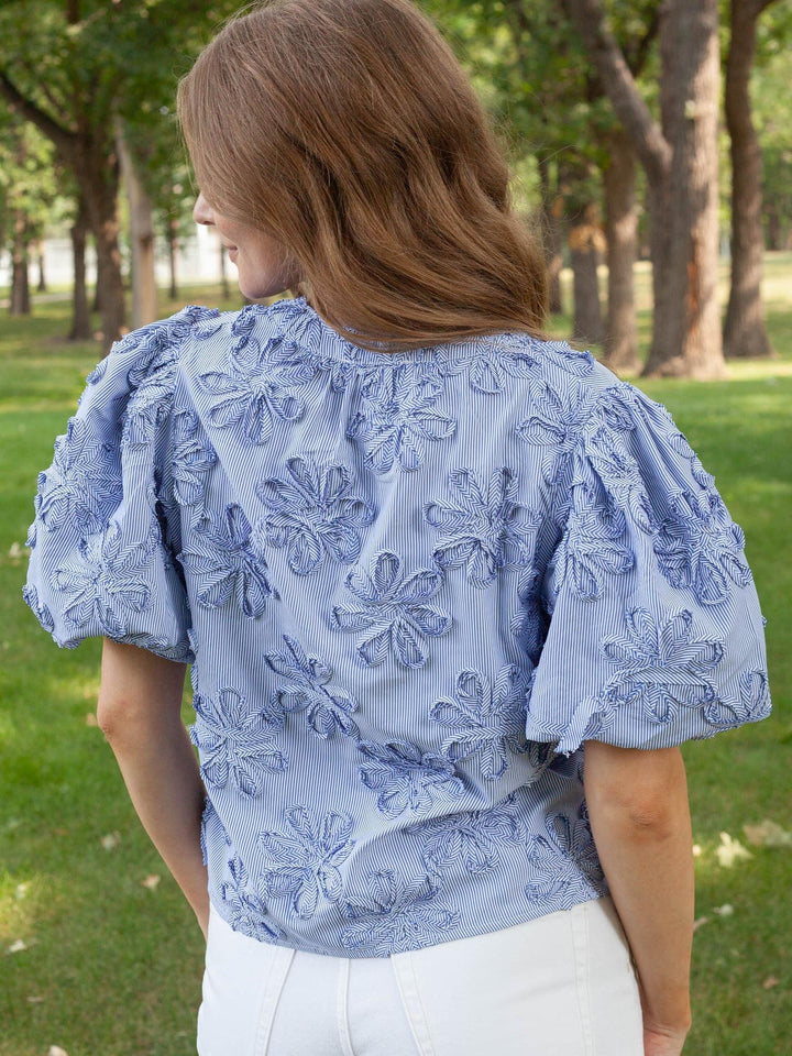 A woman viewed from behind wearing a light blue puff-sleeve blouse with raised floral embroidery, paired with white pants, standing outdoors in a park with trees and grass in the background.